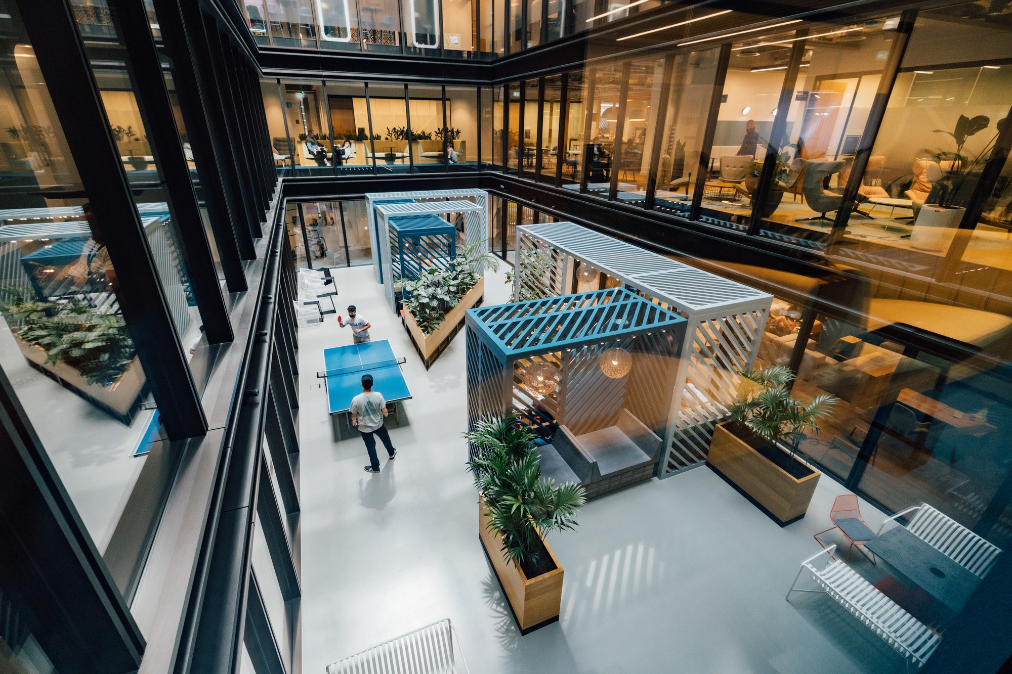 Overhead view of a modern office atrium where two people play table tennis, surrounded by plants, timber planters, blue and white slatted pergolas and glass-walled meeting spaces