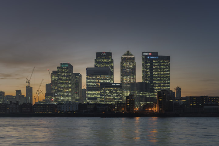The Canary Wharf skyline in London at dusk, with lit office towers reflected in the River Thames
