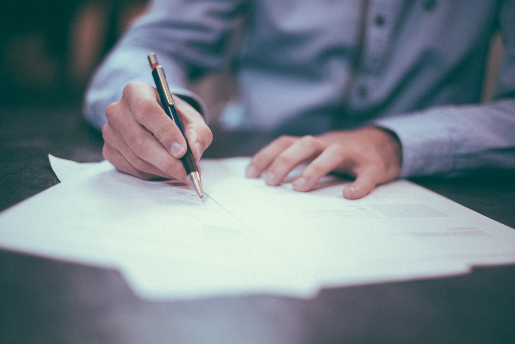 A person in a light blue shirt holding a pen over papers on a desk, about to sign a document