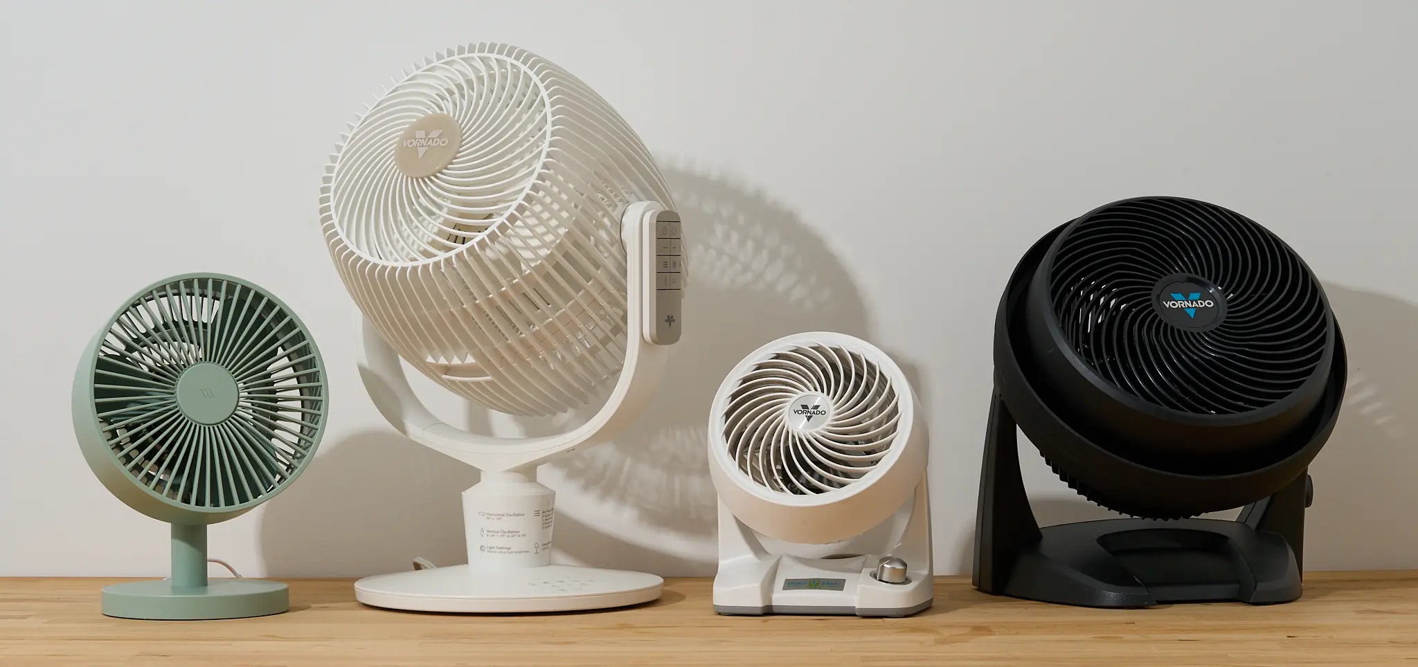 Four desk and circulator fans in a row on a wooden shelf against a white wall: a small green fan, a large white Vornado fan, a compact white Vornado fan and a black Vornado fan