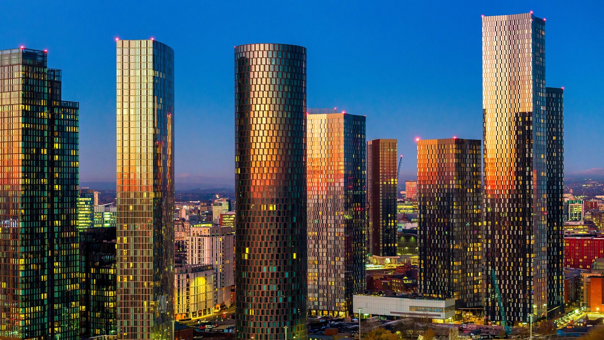 Manchester city centre skyscrapers at dusk, their glass facades glowing orange and gold against a deep blue sky